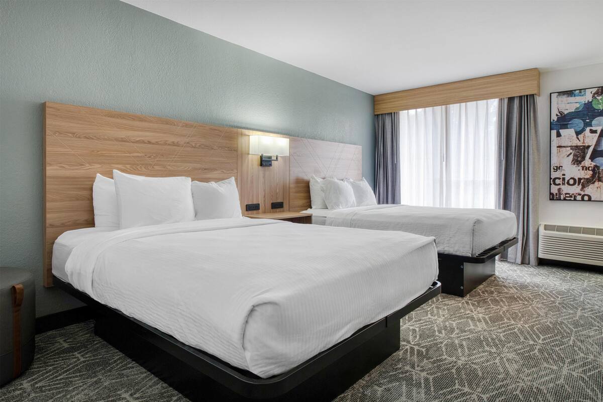 Habitación de hotel con dos camas, cabeceras, mesitas de noche, una lámpara de pared, una ventana con cortinas y un cartel en la pared.