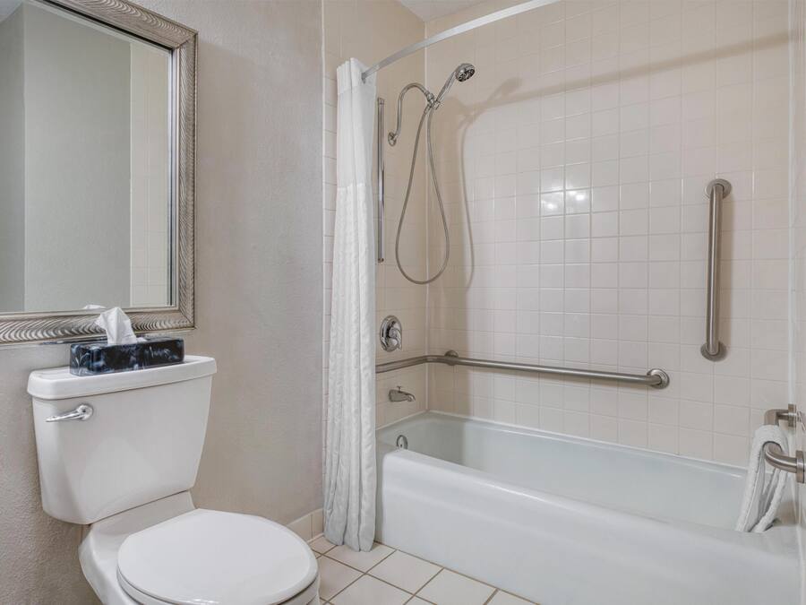 Un baño con inodoro, lavamanos, espejo, ducha con barra de apoyo y bañera.