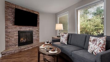 Una sala de estar moderna con chimenea de piedra, un televisor de pantalla plana montado encima, un sofá gris con cojines estampados, una mesa de centro redonda con un frutero y copas de vino, y una ventana con persianas.