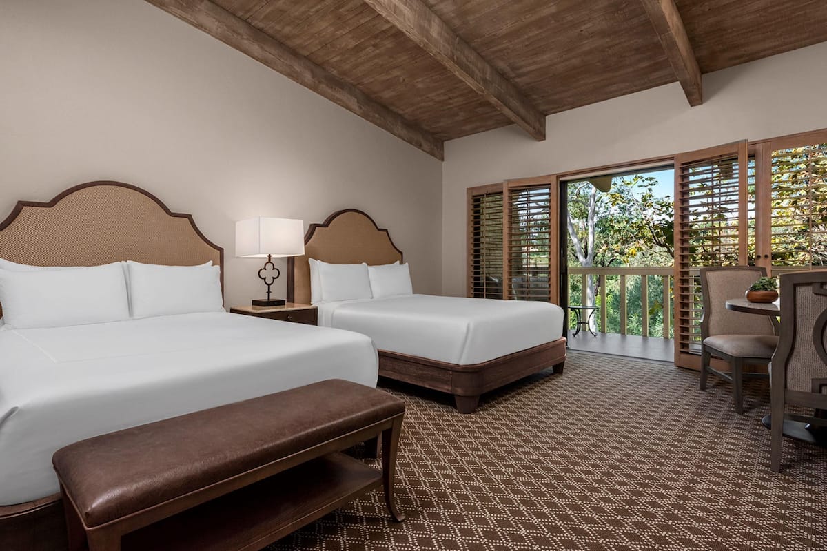 A hotel room with two beds, a bench, a table, and a balcony with a view of trees.