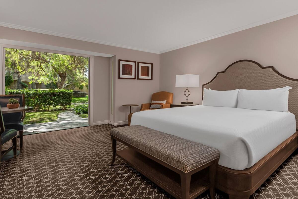 A hotel room with a large bed, a bench, a chair, a desk, and a view of a garden through a glass door.