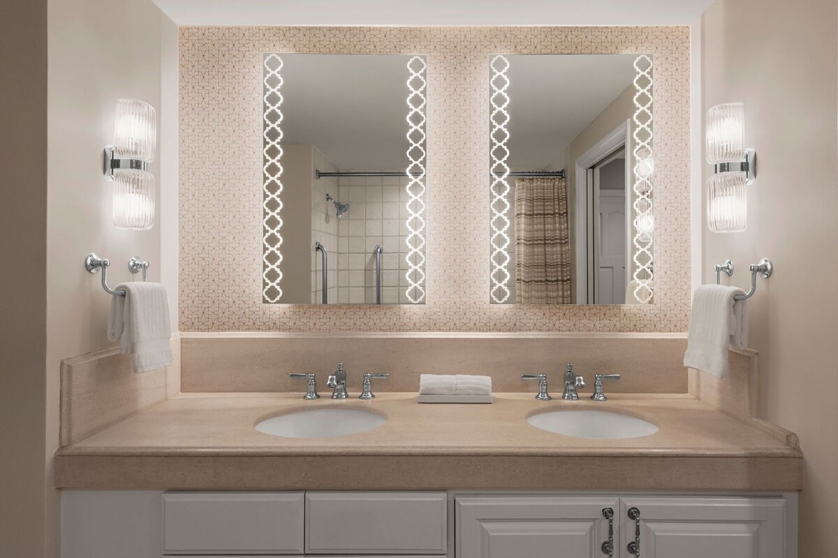 A bathroom with two sinks, a large mirror with decorative lights, and a shower visible in the reflection.