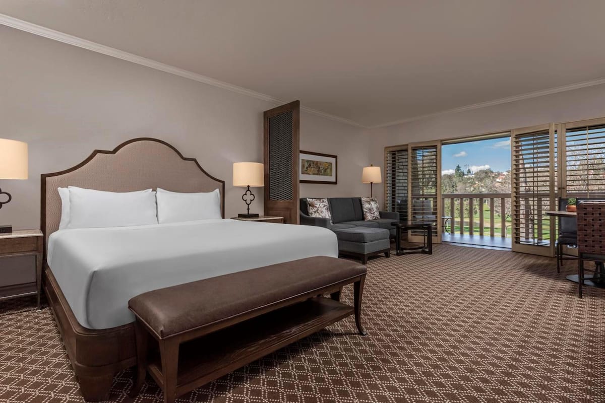 A hotel room with a large bed, a bench, two chairs, a small table, and a balcony with a view.