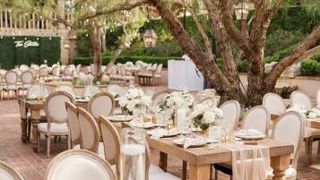 Un área de comedor al aire libre con mesas de madera y sillas blancas, ubicada bajo árboles grandes.