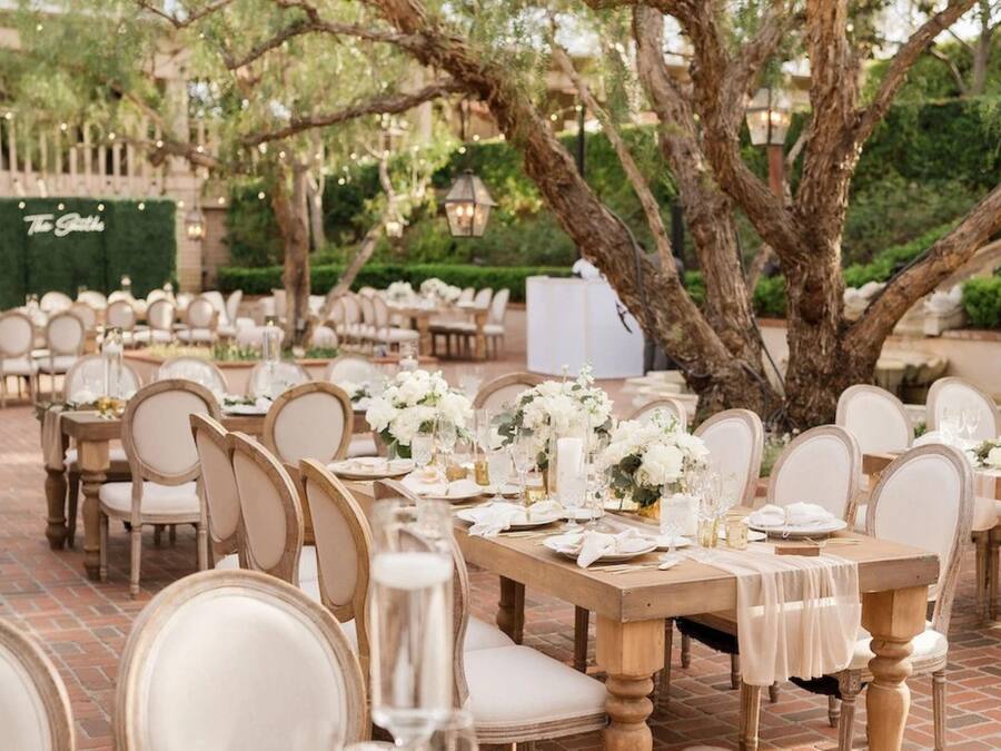 Un área de comedor al aire libre con mesas de madera y sillas blancas, ubicada bajo árboles grandes.