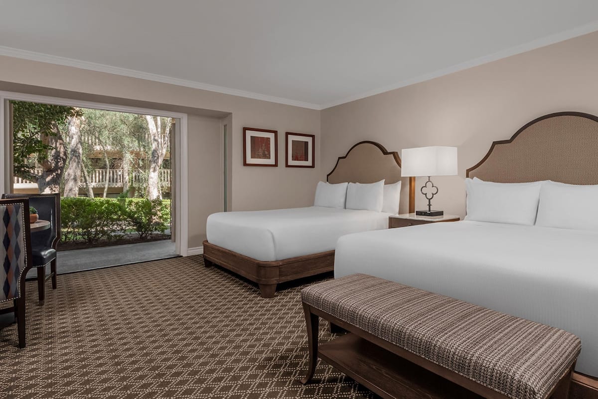 A hotel room with two beds, a bench, a desk, and a view of the outdoors.