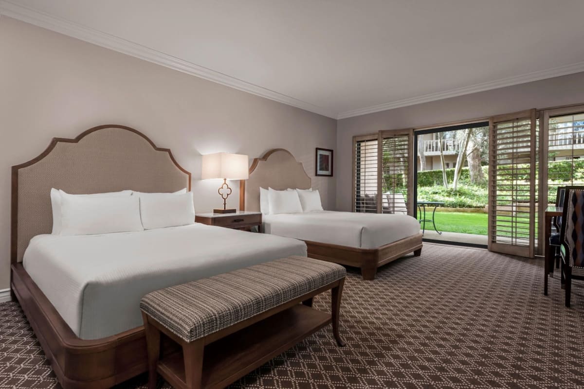 A hotel room with two beds, a bench, a nightstand, a lamp, and a view of the outdoors through large windows.