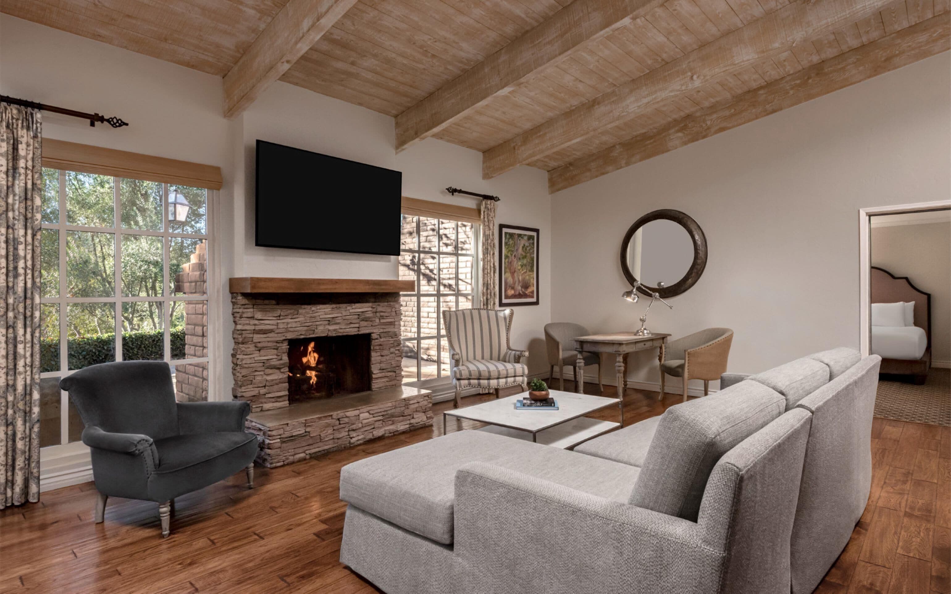 A living room with a stone fireplace, a flat-screen TV mounted above it, a grey sofa, a striped armchair, and a round mirror on the wall.