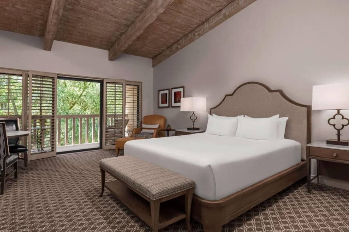 A bedroom with a large bed, a desk, a chair, a bench, and a window with a view of trees.