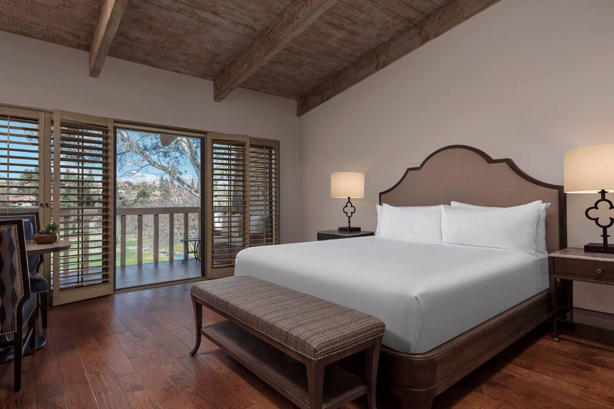 A bedroom with a large bed, two bedside tables with lamps, a bench, and a view through a wooden door.