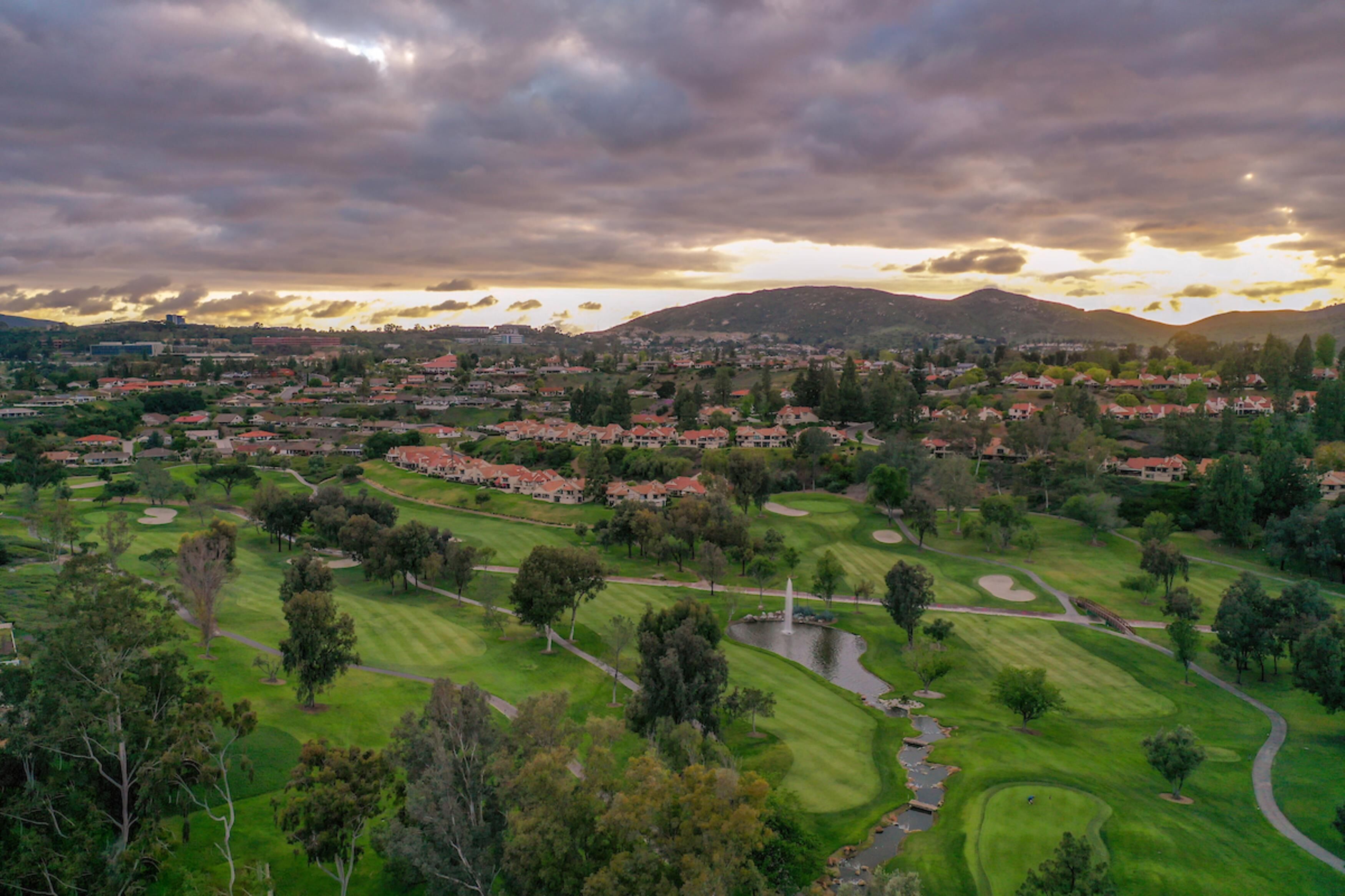 A golf course with a pond, surrounded by residential houses and hills in the background.