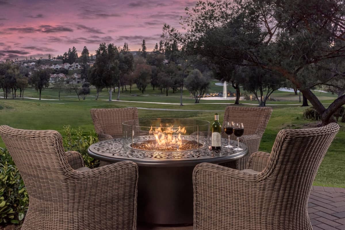 A round outdoor dining table with wicker chairs, a fire pit, and a bottle of wine.