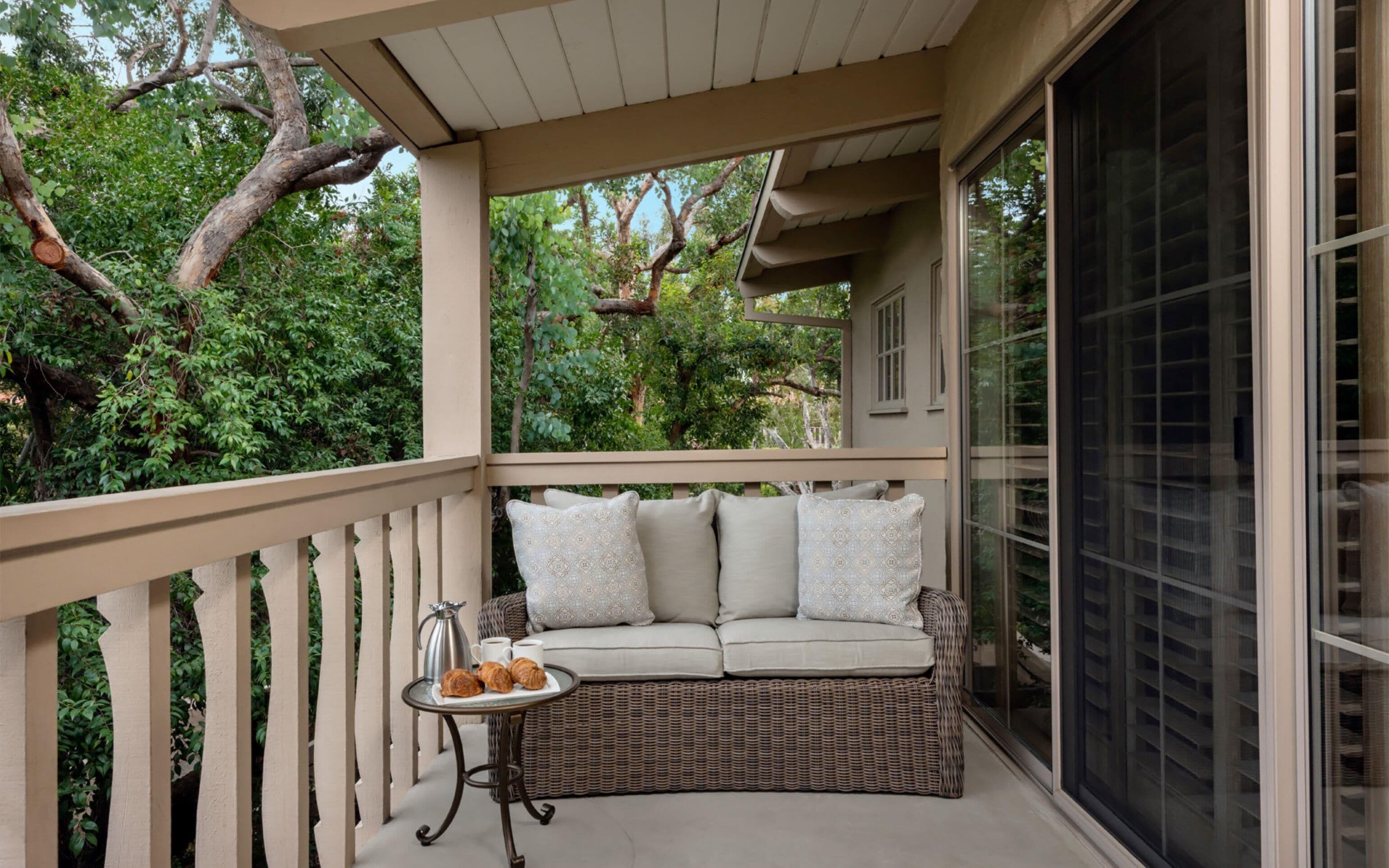 A covered balcony with a wicker sofa, a small table with croissants and coffee, and a view of lush greenery.