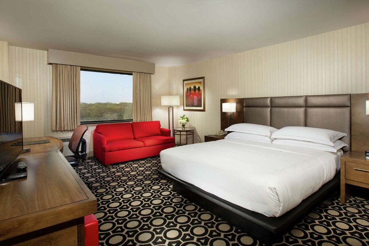A hotel room with a large bed, a red armchair, a desk with a TV, and a window with curtains.