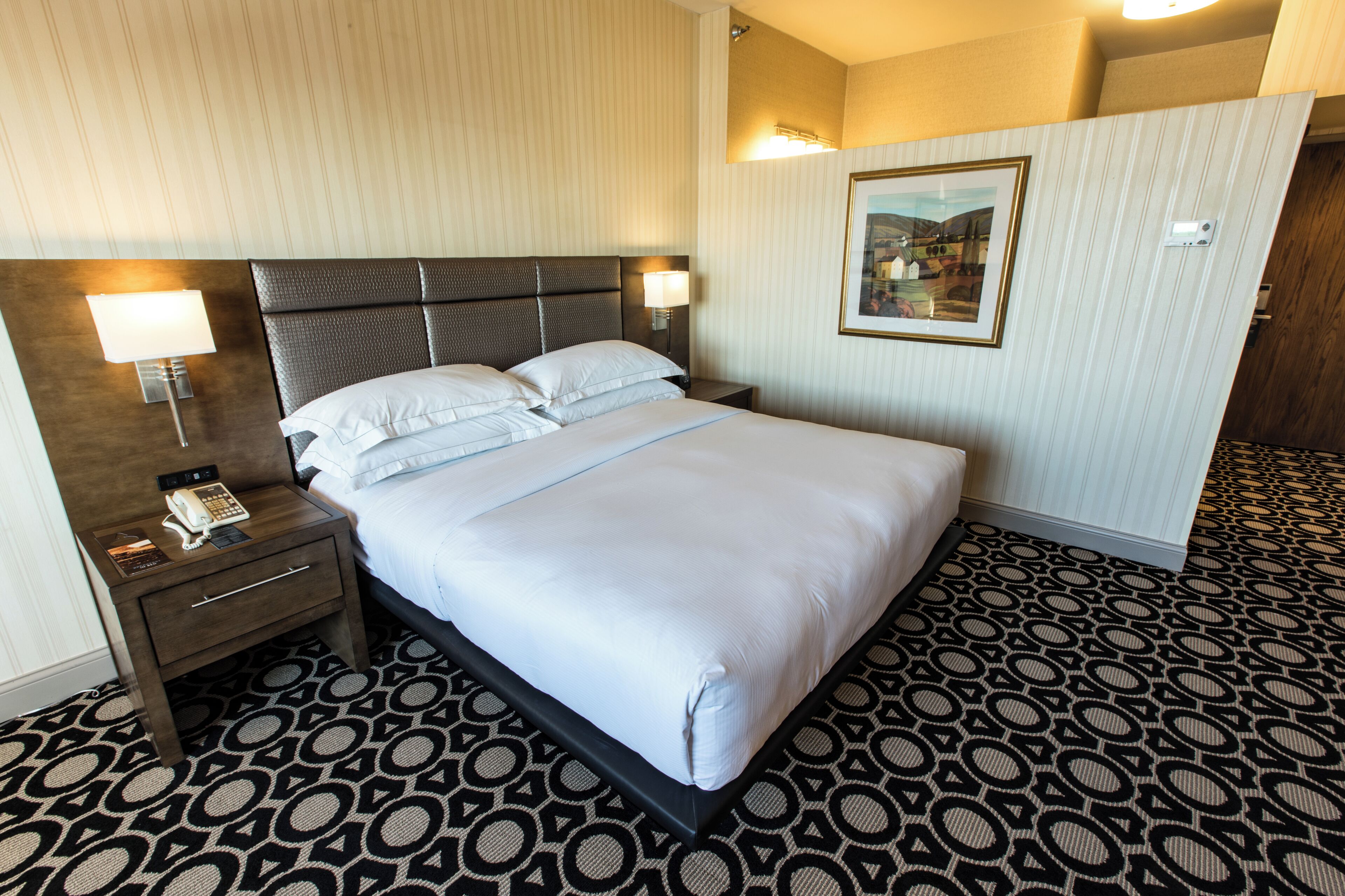 A hotel room with a large bed, a bedside table with a lamp, a phone, and a framed picture on the wall.