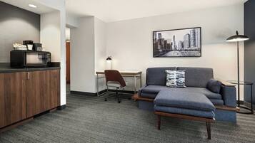 Une chambre d’hôtel moderne équipée d’une kitchenette, d’un canapé gris, d’un bureau et d’une œuvre d’art représentant un paysage urbain accrochée au mur.