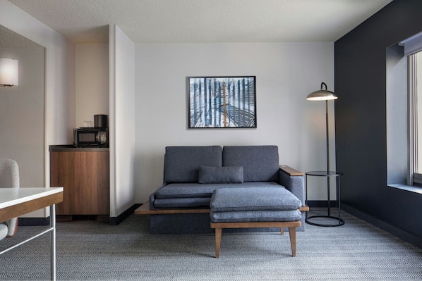 A modern room with a grey sectional sofa, a small wooden table, a floor lamp, and a framed picture of a bridge. - Courtyard by Marriott Chicago Downtown River North (Chicago)