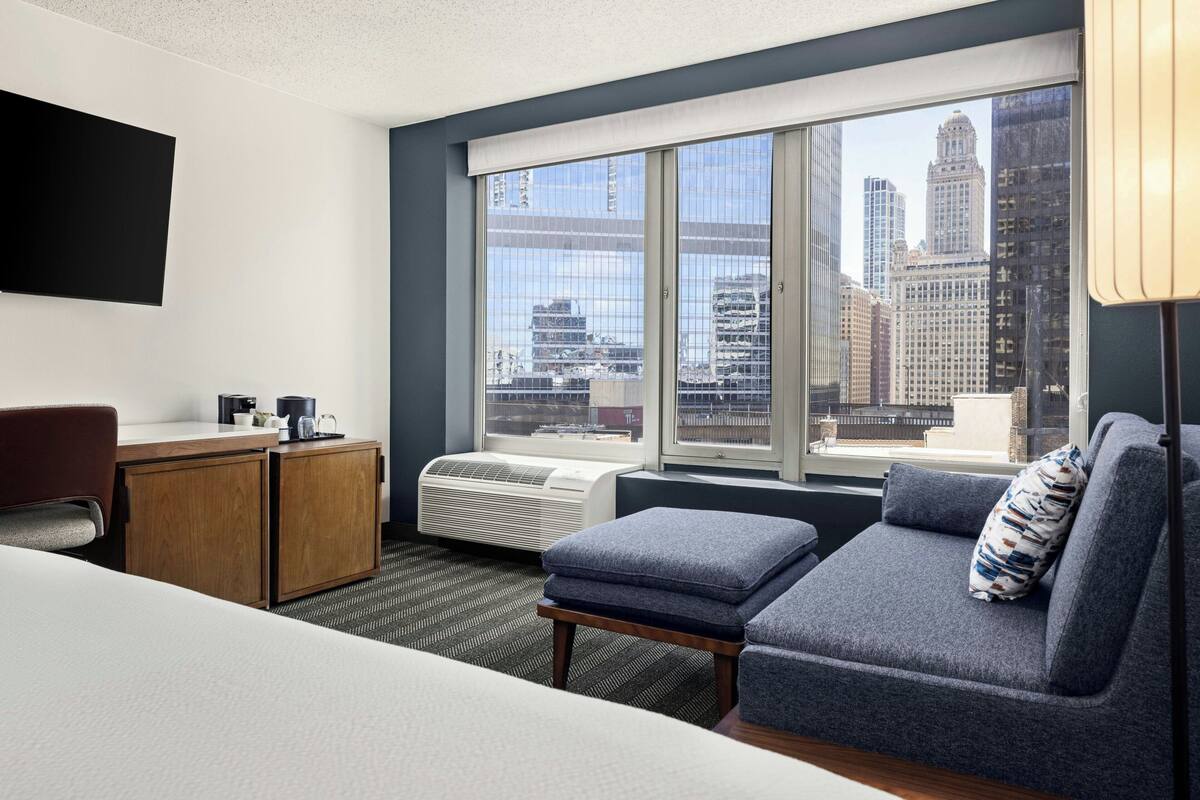 Camera d'hotel con un letto, una scrivania, una sedia, un divano e un'ampia finestra con vista sulla città.