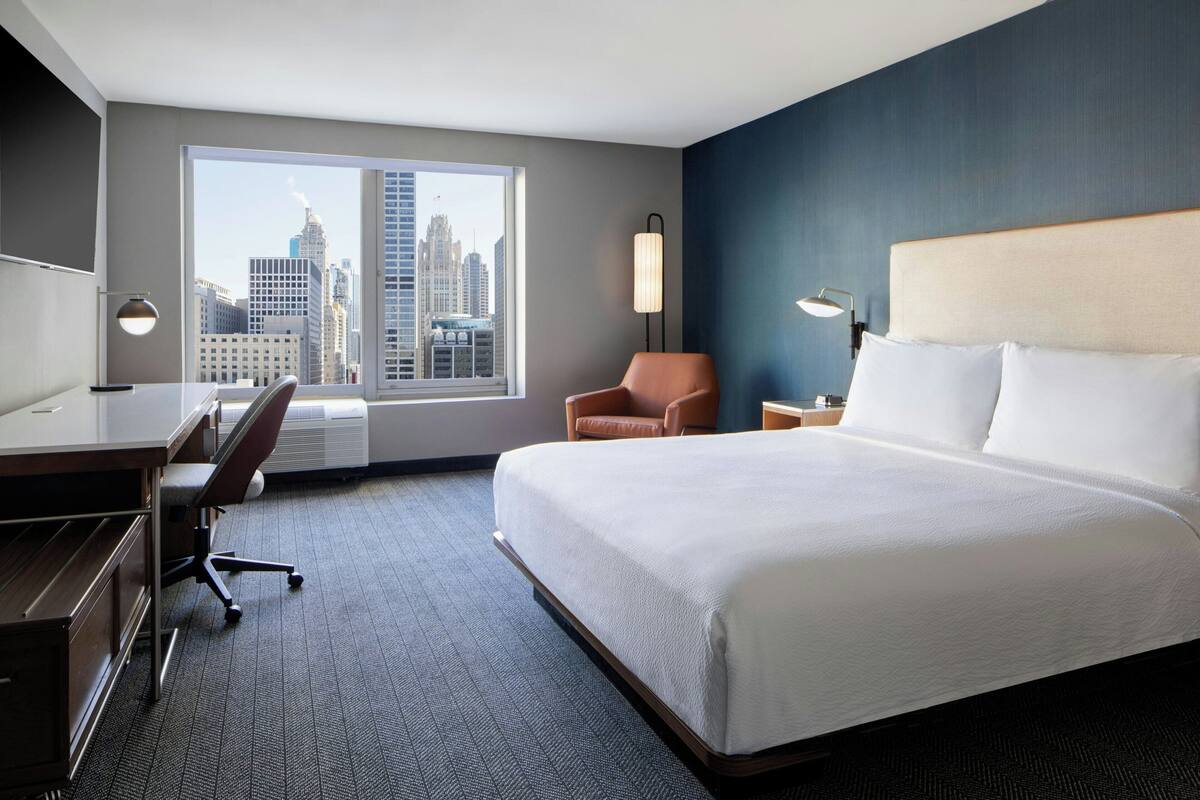 Una camera d'hotel con un letto grande, una scrivania con una sedia, una lampada e vista sullo skyline cittadino attraverso un'ampia finestra.