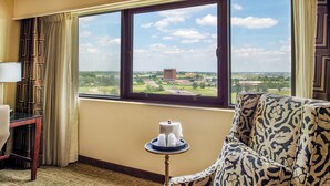 A hotel room with a large window offering a view of a distant building and landscape. There is a patterned sofa, a small table with a cake, and a lamp on a desk.