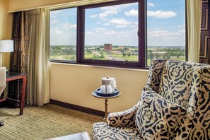 A hotel room with a large window offering a view of a distant building and landscape. There is a patterned sofa, a small table with a cake, and a lamp on a desk. - Crowne Plaza St. Louis Airport by IHG (Bridgeton)
