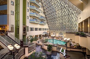 Un vestíbulo de hotel de varios niveles con una piscina central, múltiples balcones y un techo de vidrio geométrico distintivo.