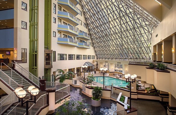 Un vestíbulo de hotel de varios niveles con una piscina central, múltiples balcones y un techo de vidrio geométrico distintivo.