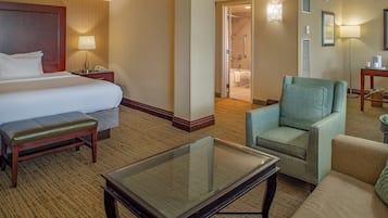 A hotel room with a large bed, a bench, a glass-top table, and a seating area with a chair and sofa.