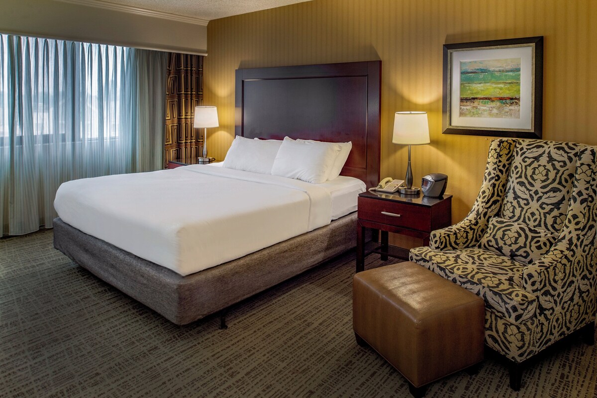 A hotel room with a large bed, a nightstand with a lamp, a patterned armchair, and a framed picture on the wall.