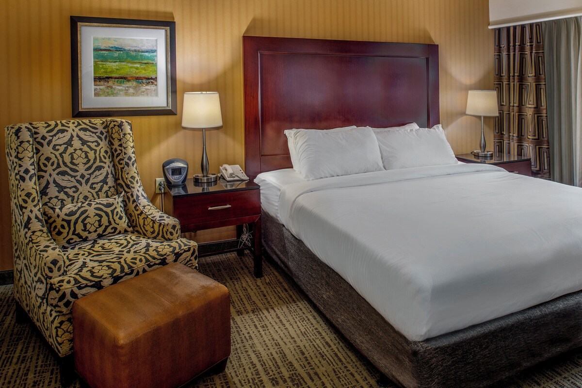 A hotel room with a large bed, a patterned armchair, a wooden nightstand, and a framed painting on the wall.