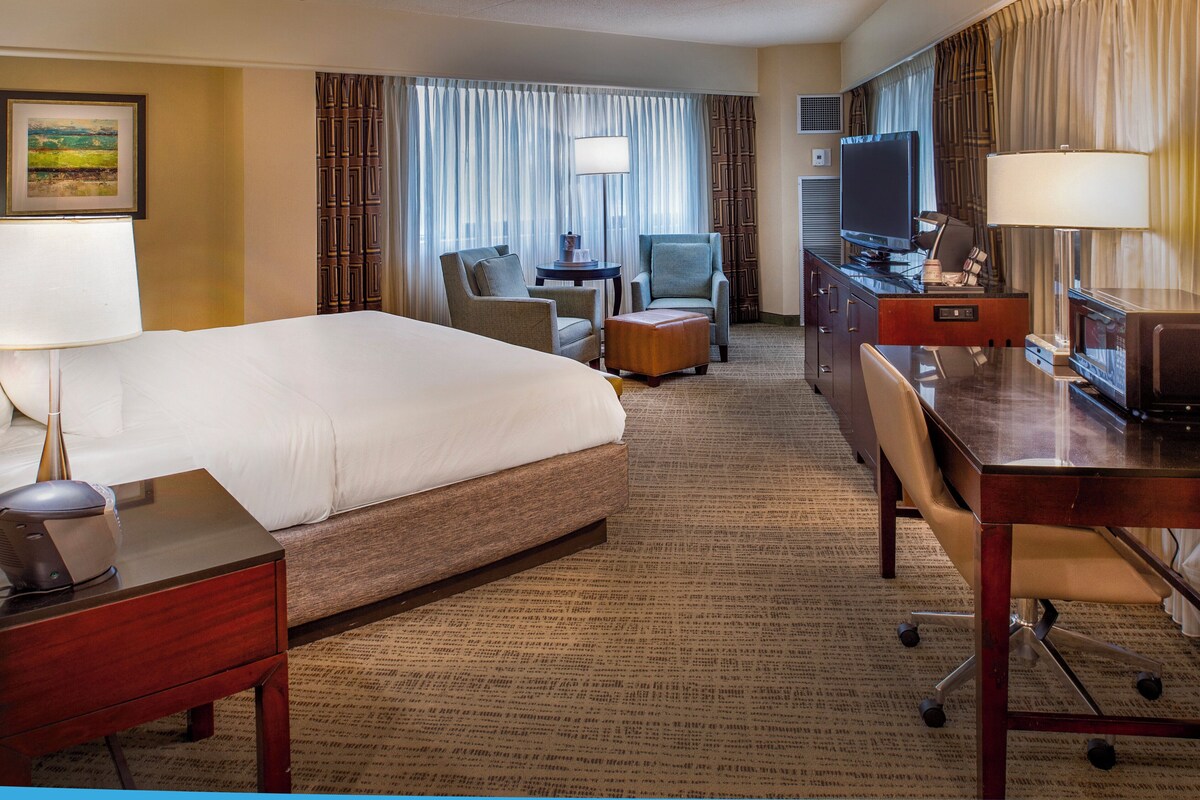 A hotel room with a large bed, a desk with a computer, a chair, a small table, and a TV.