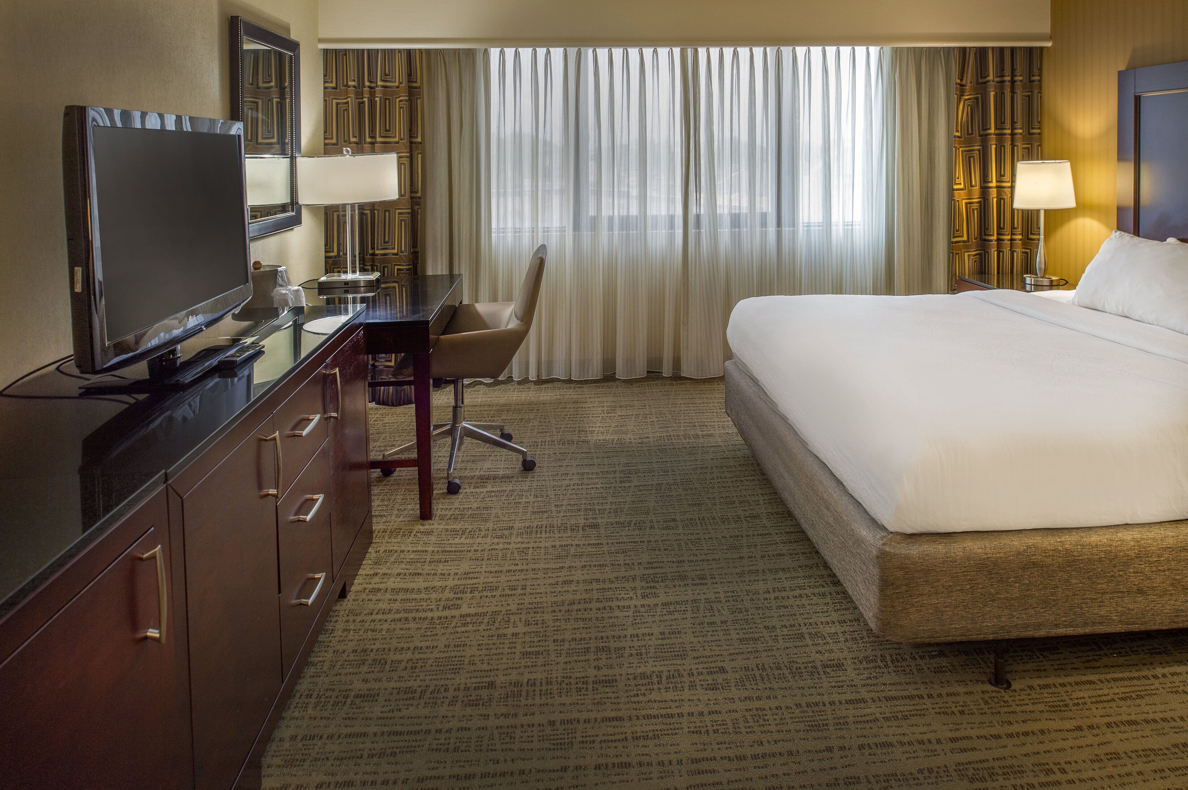 A hotel room with a large bed, a desk with a computer, a television, and a window with sheer curtains.