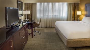 A hotel room with a large bed, a desk with a computer, a television, and a window with sheer curtains.