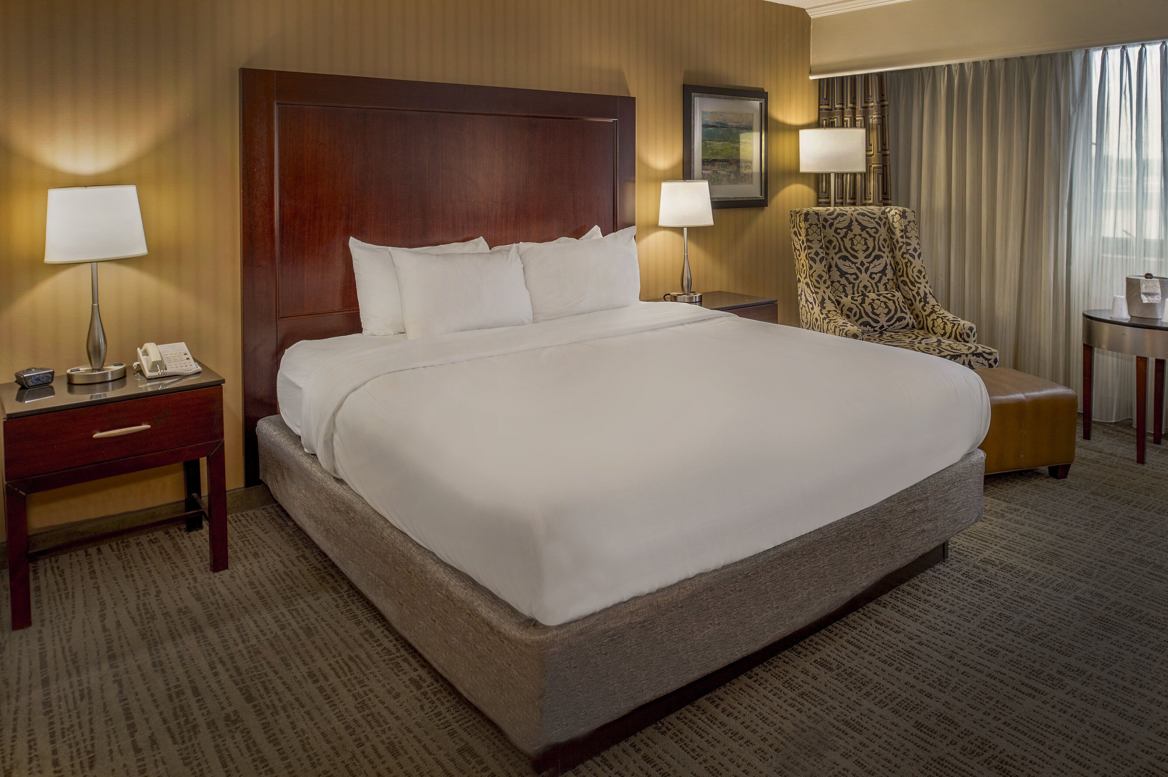 A large bed with white linens, a wooden headboard, two bedside tables with lamps, a patterned armchair, and a small table with a lamp.