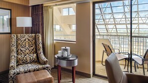 A hotel room with a patterned armchair, a small table with a lamp, and a view of a balcony with chairs.