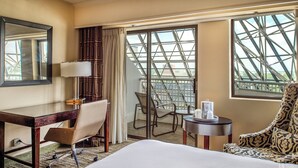 A hotel room with a bed, a desk, a chair, a small table, and a large window with a view of a building structure.