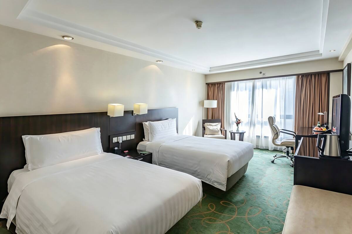 A hotel room with two beds, a desk with a chair, a TV, and a large window with curtains.