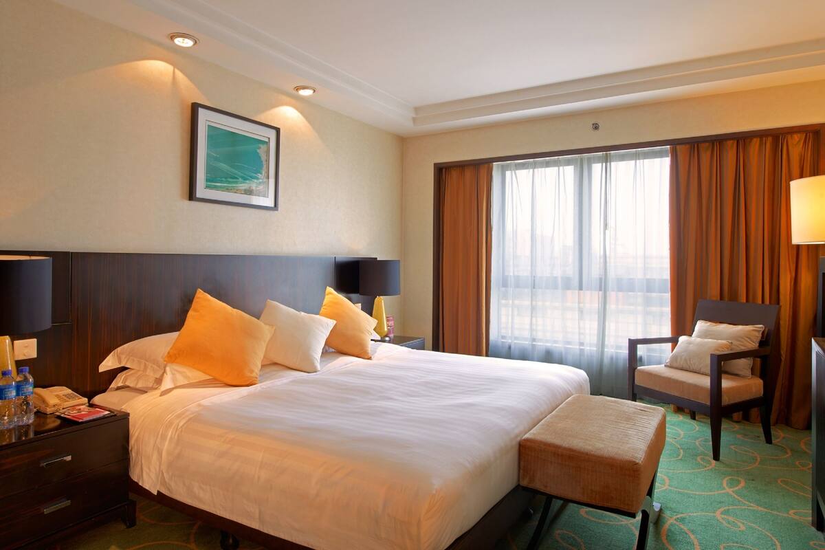 A hotel room with a large bed, two bedside lamps, a nightstand with a phone and water bottles, a desk with a chair, and a window with curtains.