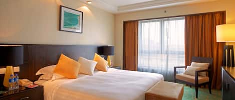 Habitación de hotel con una cama grande, dos lámparas de noche, una mesita de noche con un teléfono y botellas de agua, un escritorio con una silla y una ventana con cortinas.