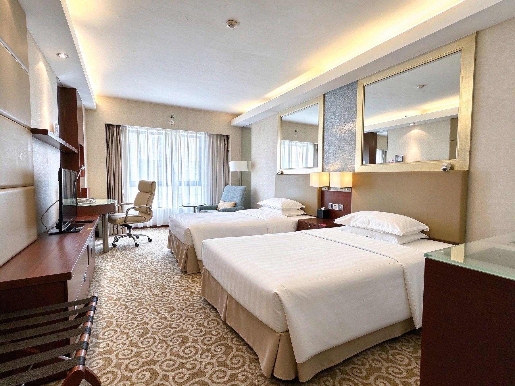 A hotel room with two beds, a desk, a chair, a desk lamp, and a large mirror.