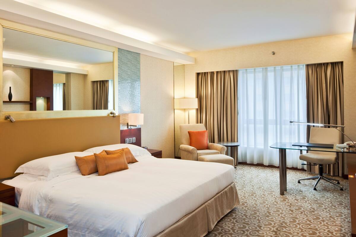 A hotel room with a large bed, a desk, a chair, a lamp, a mirror, and a window with curtains.