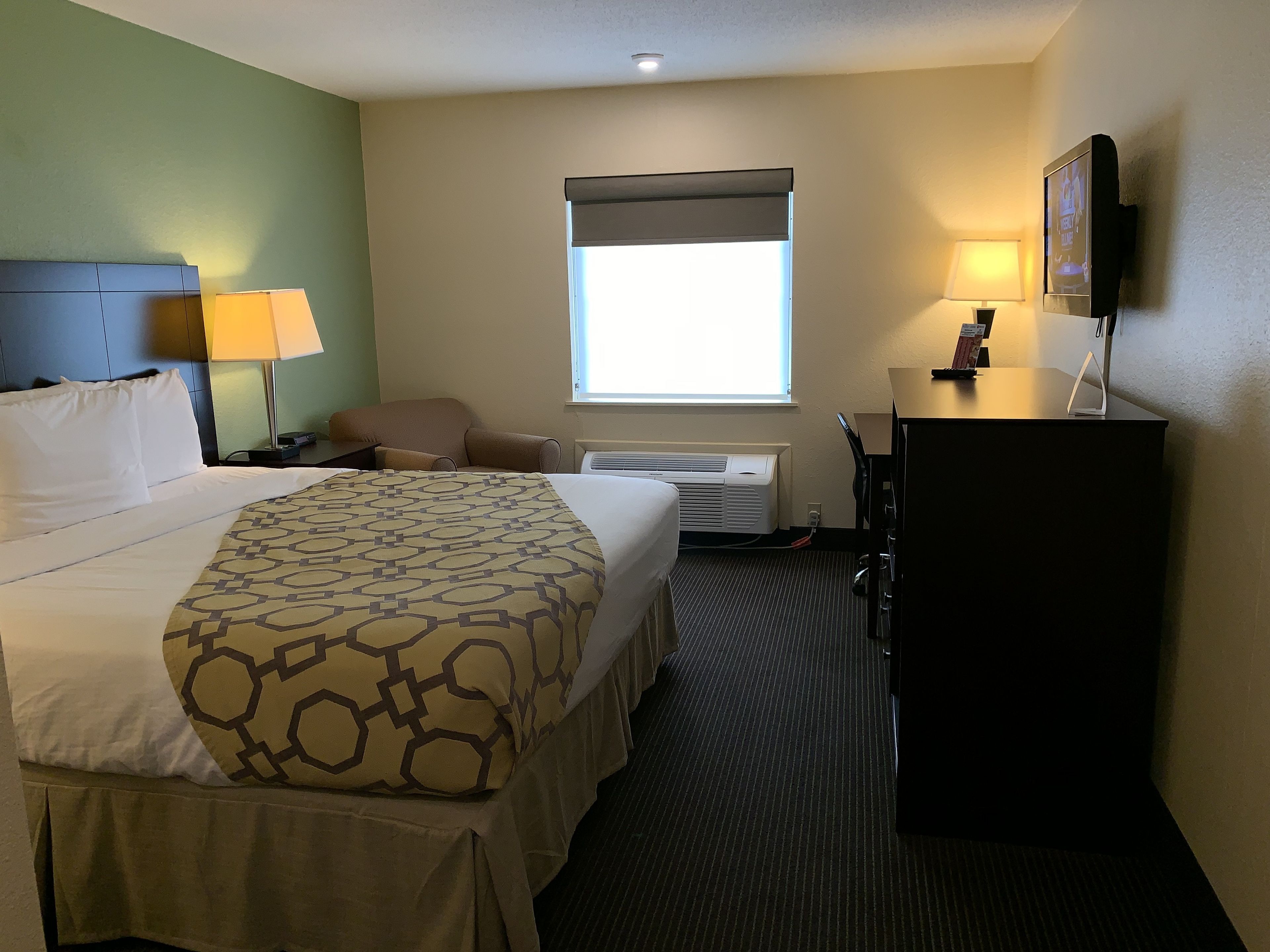 A hotel room with a large bed, a desk, a chair, a TV, and a window with blinds.