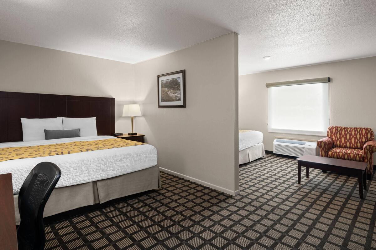 Ein Hotelzimmer mit einem großen Bett, einem Schreibtisch, einem Sessel, einem kleinen Tisch, einer Lampe, einem Bild an der Wand und einem Fenster mit Jalousie.