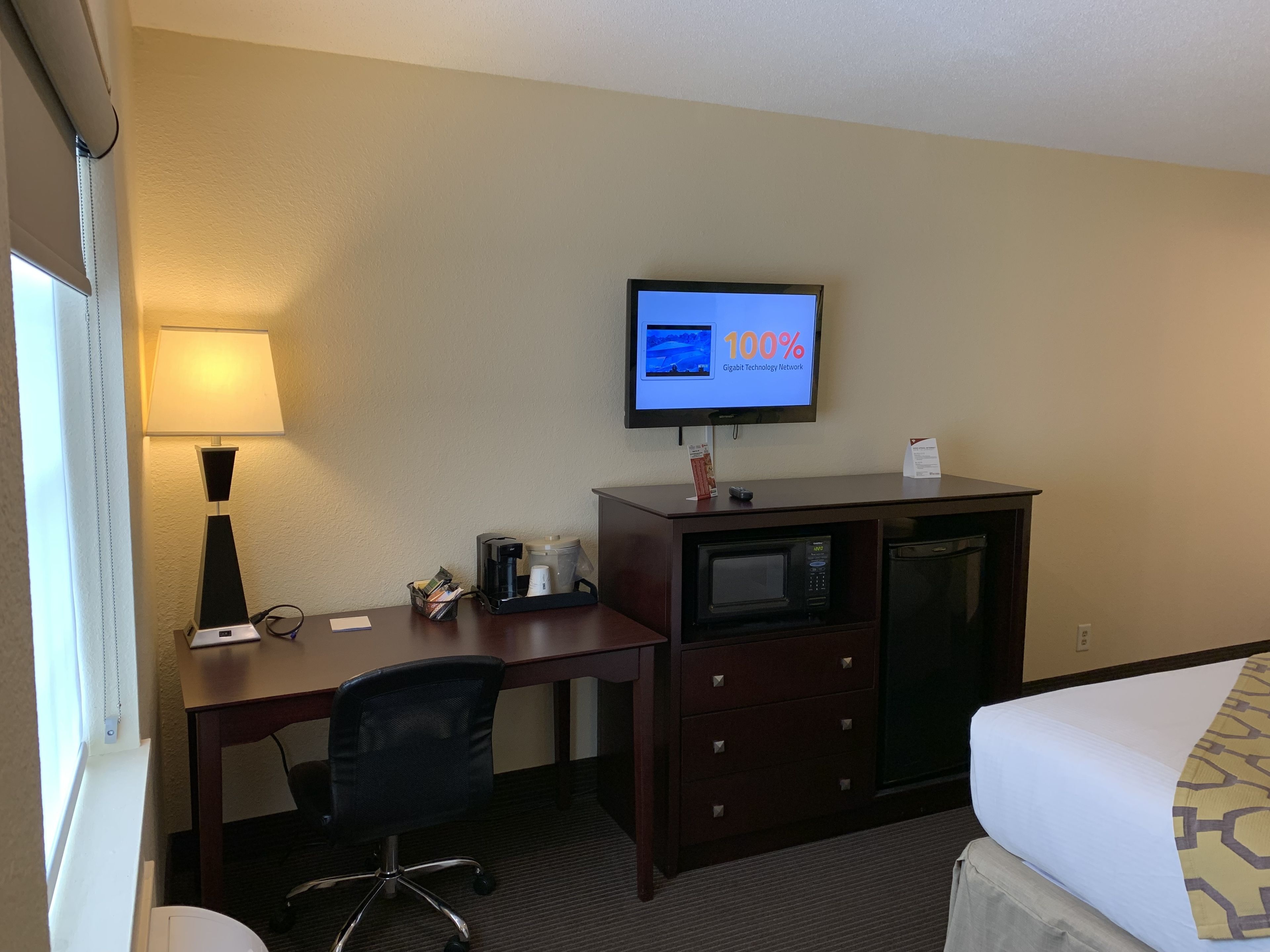 Habitación de hotel con cama, escritorio con lámpara, televisión mostrando un anuncio y cafetera, además de un microondas.