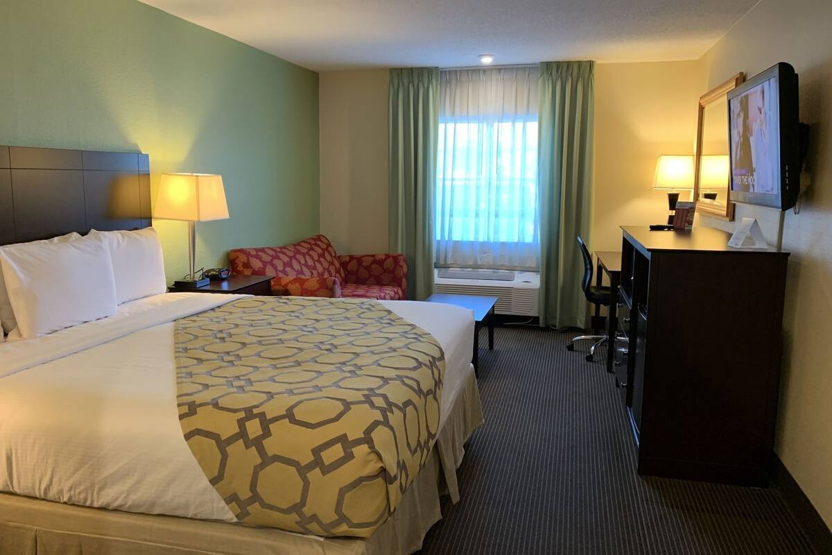 Ein Hotelzimmer mit einem großen Bett, einem Schreibtisch, einem Stuhl, einem Fernseher und einem Fenster mit Vorhängen.