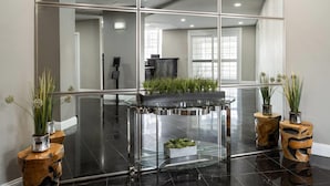 Een moderne lobby met een glazen tafel, twee houten bijzettafeltjes met planten, en een grote spiegel die het interieur reflecteert.