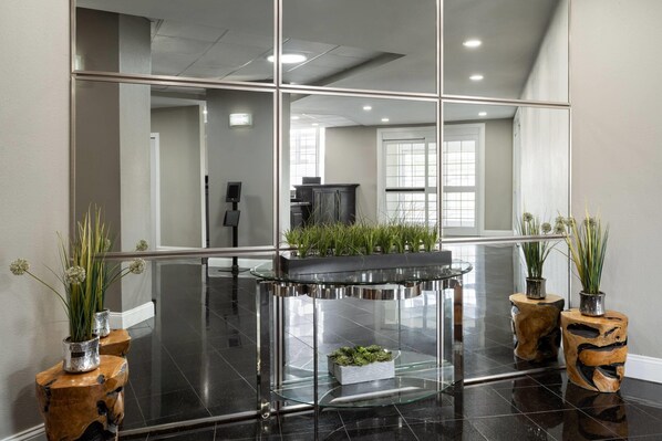 A modern lobby with a glass table, two wooden side tables with plants, and a large mirror reflecting the interior.