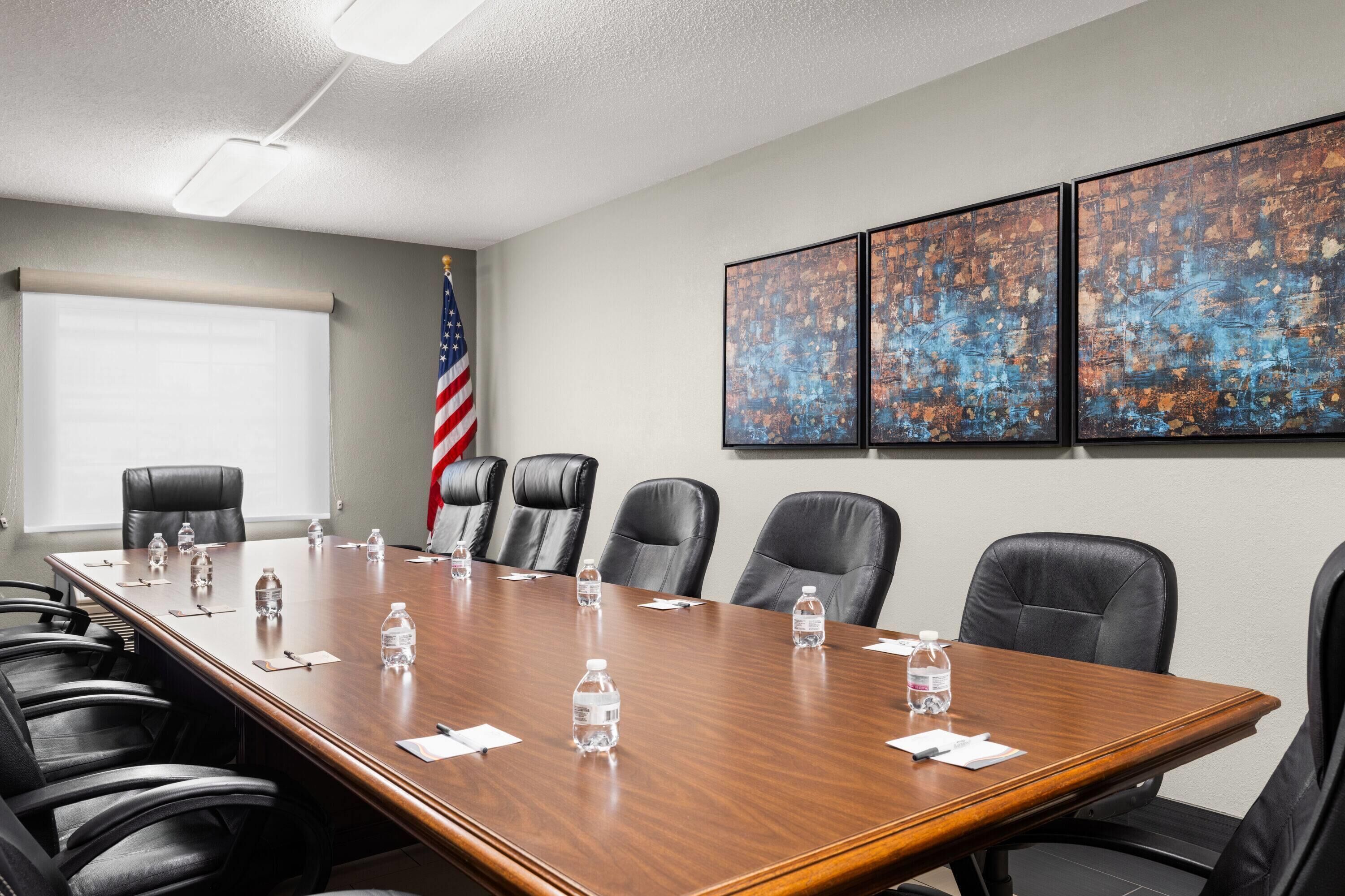 Une salle de conférence avec une longue table, des fauteuils en cuir noir et un drapeau américain. On trouve des bouteilles d’eau sur la table et trois tableaux abstraits au mur.