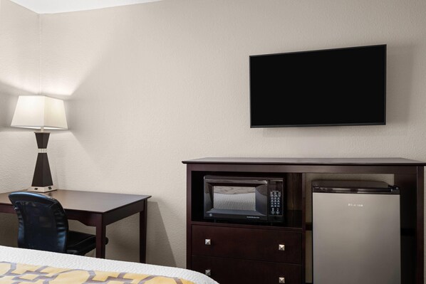 A hotel room with a bed, a desk with a chair, a lamp, a flat-screen TV, a microwave, and a refrigerator.