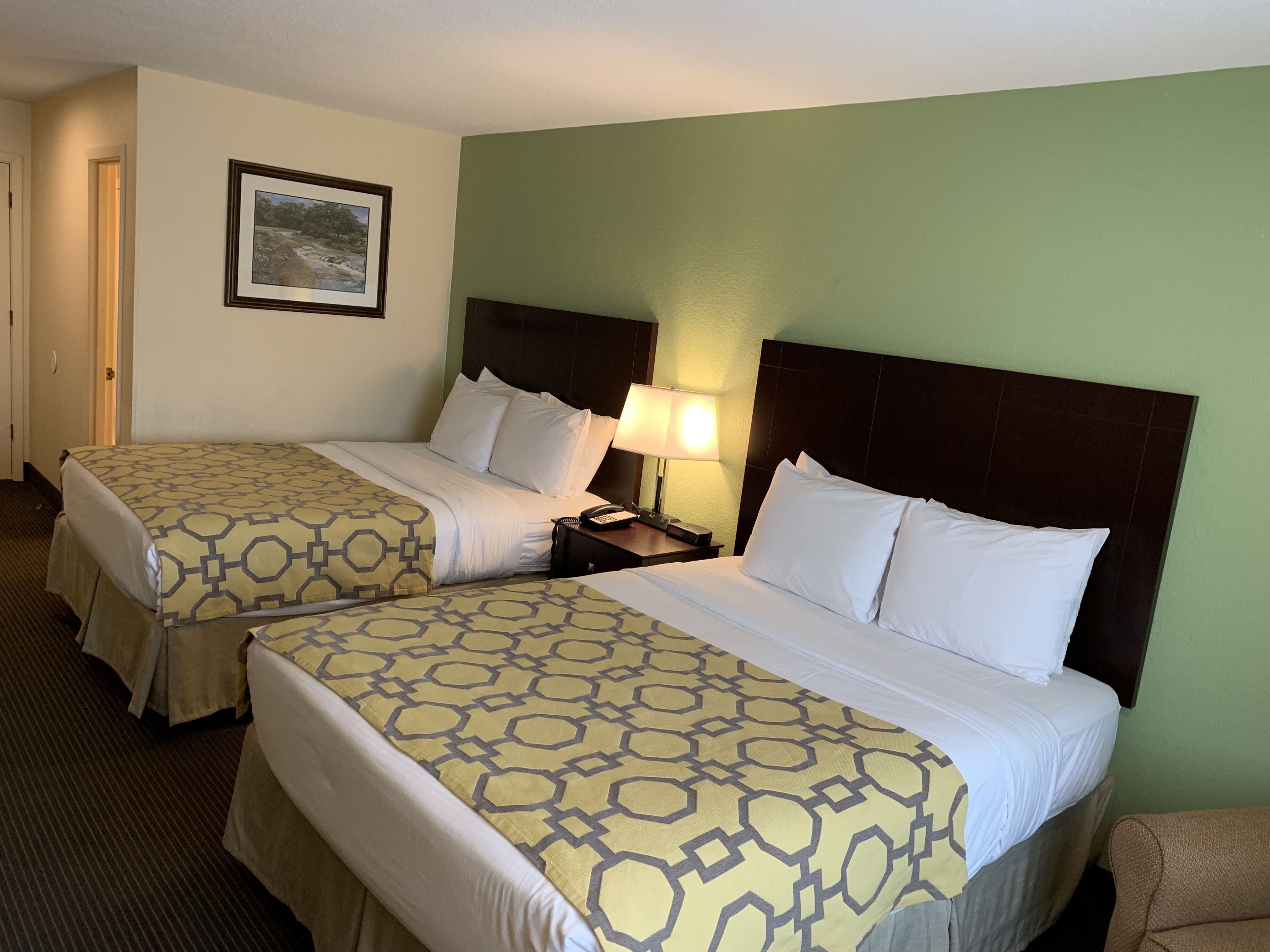 A hotel room with two beds, a nightstand with a lamp, a framed picture on the wall, and a green accent wall.